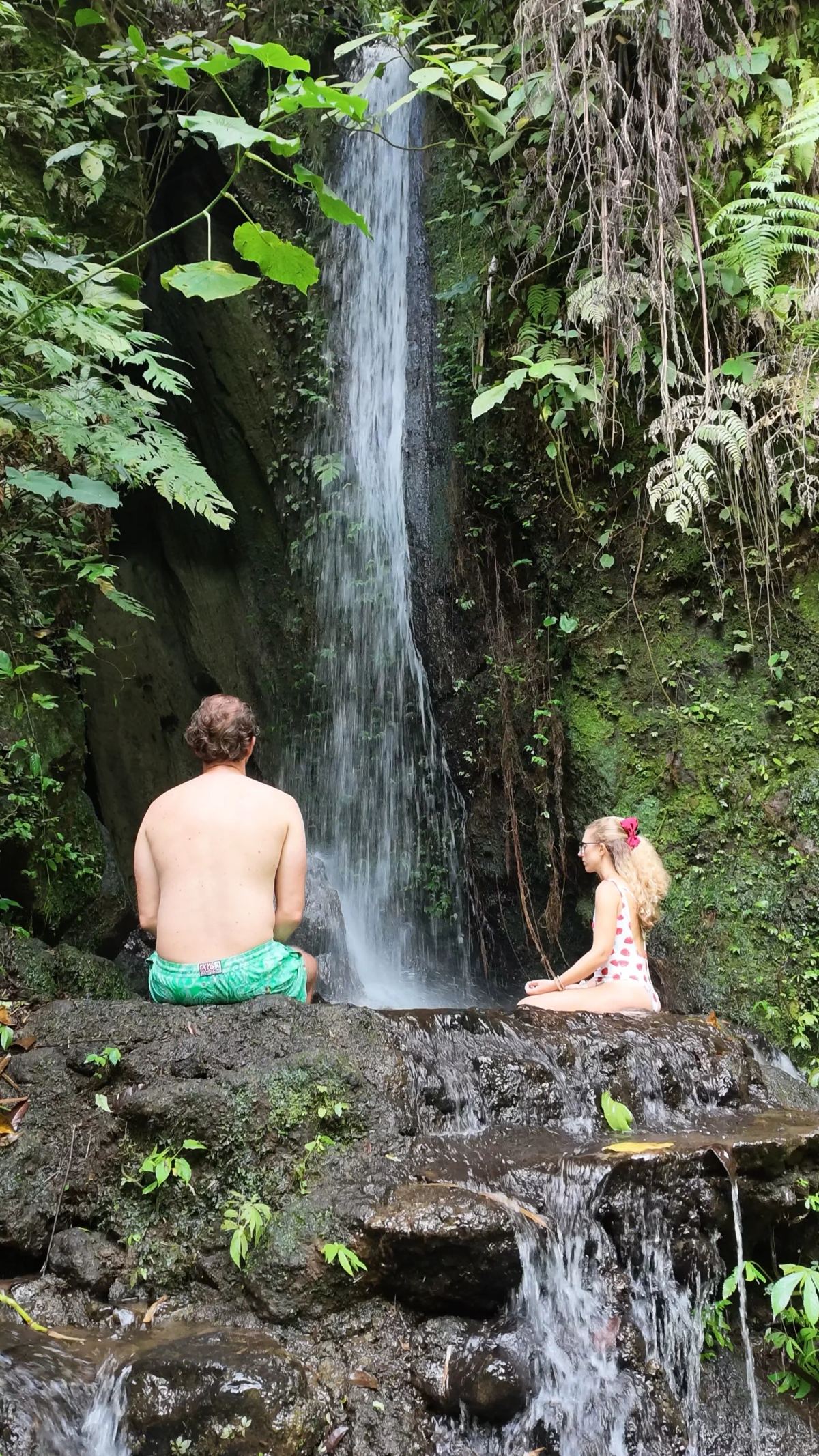Due persone sedute in acqua davanti a una piccola cascata tra le rocce.